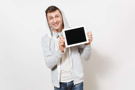 Young handsome smiling man in t-shirt and light sweatshirt with hood with headphones holds tablet pc computer with blank screen for copy space isolated on white background. Concept of technologyの写真素材