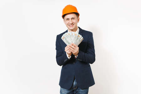 Young businessman in dark suit, protective construction helmet holding bundle of dollars, cash money and covering face isolated on white background. Male worker for advertisement. Business conceptの写真素材