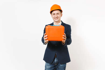 Young smiling businessman in dark suit, protective hardhat holding case with instruments or toolbox...