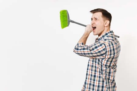 Young combative angry housekeeper man in checkered shirt holding and sweeping with green broom isolated on white background. Male doing house chores. Copy space for advertisement. Cleanliness conceptの写真素材