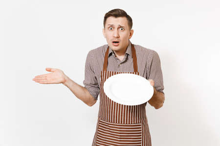 Young man chef or waiter in striped brown apron shirt holding white round empty clear plate isolated...