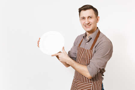 Young man chef or waiter in striped brown apron shirt holding white round empty clear plate isolated on white background. Male housekeeper or houseworker. Domestic worker copy space for advertisementの写真素材
