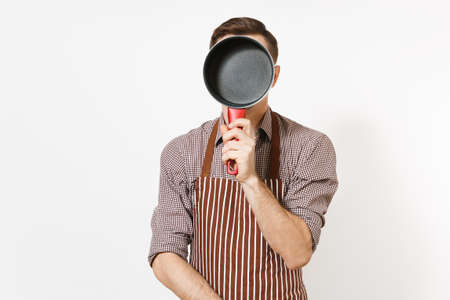 Young fun crazy man chef or waiter in striped brown apron, shirt holding red empty stewpan on face isolated on white background. Male housekeeper or houseworker. Kitchenware and cuisine conceptの写真素材