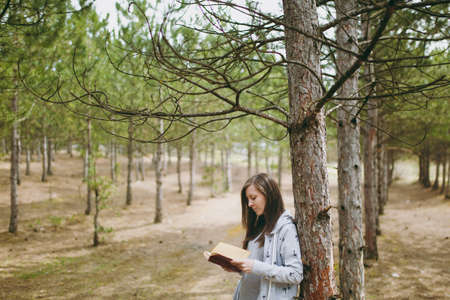 Young calm beautiful woman in casual clothes leaning on tree studying reading book in big city park...