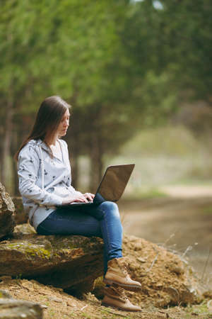 Young serious successful smart business woman or student in casual clothes sitting on stone using laptop in city park or forest working outdoors on green blurred background. Mobile Office conceptの写真素材