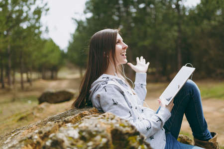 Young fun smiling beautiful woman in casual clothes with earphones sitting on stone using tablet pc...