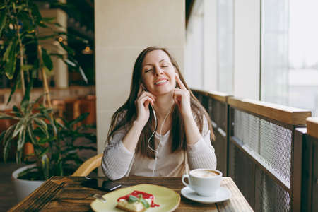 Young woman sitting in coffee shop at table with cup of cappuccino, cake, relax in restaurant during...