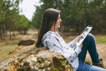 Young smiling beautiful woman in casual clothes with earphones sitting on stone using tablet pc...