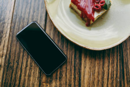 Close up of cake with raspberry sauce on plate, cup of cappuccino, mobile phone with blank empty screen lying on wooden table in coffee shop, cafe, restaurant. Overhead top view. Lifestyle conceptの写真素材