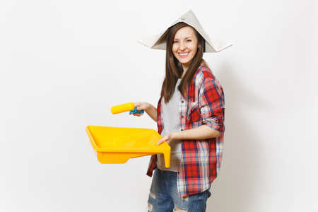 Young smiling beautiful woman in newspaper hat holding roller for wall painting and paint tray isolated on white background. Instruments, tools for renovation apartment room. Repair home conceptの写真素材