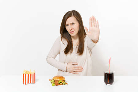 Illness woman put hand on pain abdomen, stomach-ache at table with burger, french fries, cola in glass bottle isolated on white background. Proper nutrition or American classic fast food. Copy spaceの写真素材