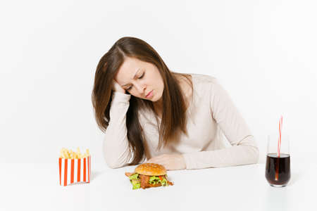 Sad tired woman put hand on head sitting at table with burger, french fries, cola in glass bottle isolated on white background. Proper nutrition or American classic fast food. Area with copy spaceの写真素材