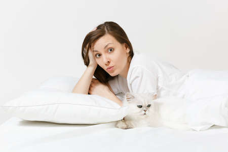 Calm young woman lying in bed with white cute Persian silver chinchilla cat, sheet, pillow, blanket on white background. Beauty female spending time in room. Rest, relax good mood concept. Copy spaceの写真素材
