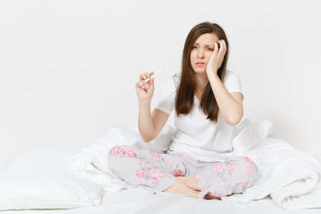 Young sick tired sad woman in pajamas sitting on bed, holding clinical thermometer with high fever temperature isolated on white background. Female feeling bad, spending time at home. Advertisingの写真素材