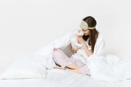 Calm young woman sitting in bed with sleep mask on eyes isolated on white background. Beauty female spending time in room with white cute Persian silver chinchilla cat. Rest, relax, good mood conceptの写真素材