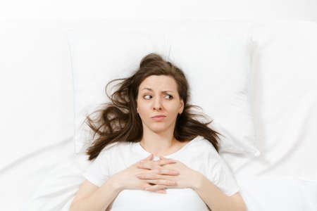 Top view of tired stressed crying young woman lying in bed with white sheet, pillow, blanket. Pensive frustrated sad upset female spending time in room. Rest, relax, bad mood concept. Copy spaceの写真素材