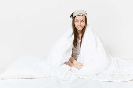 Fun tired young woman sitting in bed with sleep mask, sheet, pillow, wrapping in blanket isolated on white background. Beauty female spending time in room. Rest, relax, good mood concept. Copy spaceの写真素材
