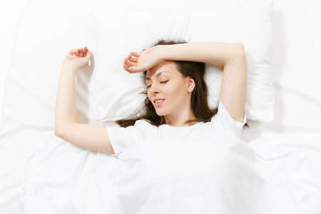 Top view of head of happy brunette young woman lying in bed with white sheet, pillow, blanket. Beautiful female spending time in room. Rest, relax, good mood concept. Copy space for advertisementの写真素材