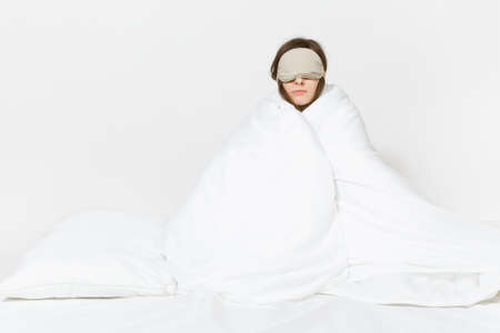 Fun tired young woman sitting in bed with sleep mask, sheet, pillow, wrapping in blanket isolated on...