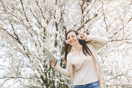 Young smiling beautiful woman in light casual clothes with headphones listening music holding tablet pc computer in city garden or park on blooming tree background.の写真素材