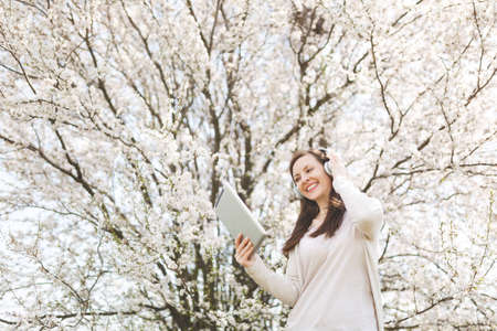 Young smiling beautiful woman in light casual clothes with headphones listening music holding tablet...
