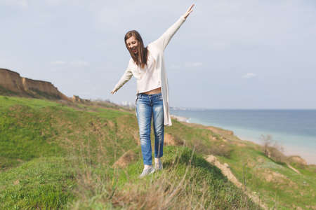 Young brunette smiling relaxed woman with closed eyes in light casual clothes spreading hands walking near water on green grass field background.の写真素材