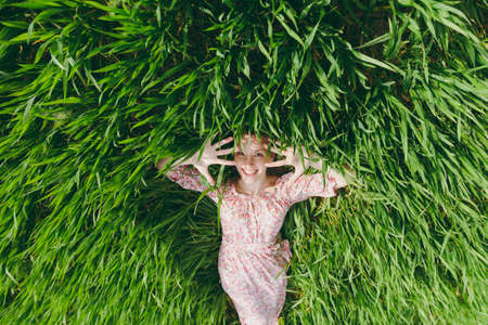 Young smiling beautiful woman in light patterned dress lying on grass showing hands looking up resting in sunny weather in field on bright green background.の写真素材