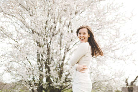 Young happy laughing beautiful woman in light casual clothes holding hands folded standing in city garden or park on blooming tree background.の写真素材