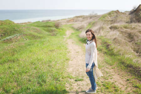Young brunette smiling beautiful woman in light casual clothes walking along path standing in sunny weather in field near water on green background.の写真素材