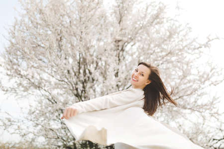 Young happy relaxed smiling beautiful woman in light casual clothes turning around standing in city garden or park on blooming tree background.の写真素材