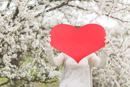 Young woman in casual clothes holding covering face with big red heart standing in city garden or...