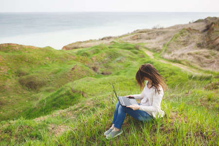 Young business woman or student in casual clothes with fluttering hair sitting on grass using laptop...