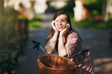 Smiling young woman in long pink floral dress stop to riding on vintage bike with basket for purchases, food or flowers outdoors, gorgeous attractive female recreation time in spring or summer parkの写真素材