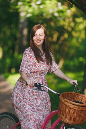 Portrait of trendy young woman in long pink floral dress stop to riding under oak arch on bike with basket for purchases food or flowers outdoors, cute female recreation time in spring or summer parkの写真素材