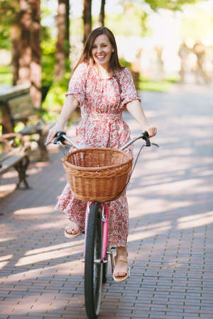 Portrait of trendy young woman in long pink floral dress riding on alley on vintage bike with basket for purchases, food or flowers outdoors, gorgeous female recreation time in spring or summer parkの写真素材