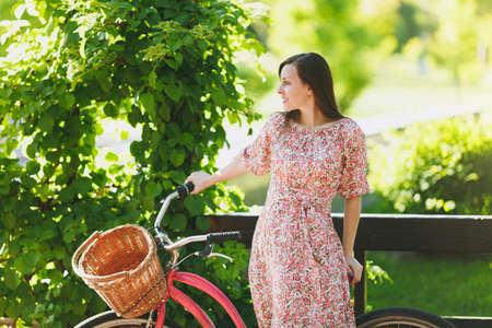 Portrait of trendy young woman in long pink floral dress stop to riding on vintage bike with basket for purchases, food or flowers outdoors, gorgeous female recreation time in spring or summer parkの写真素材