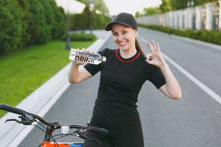 Young smiling athletic strong woman in black uniform, cap holding pointing on bottle of water, riding road on bicycle outdoors on spring or summer sunny day. Fitness, sport, healthy lifestyle conceptの写真素材