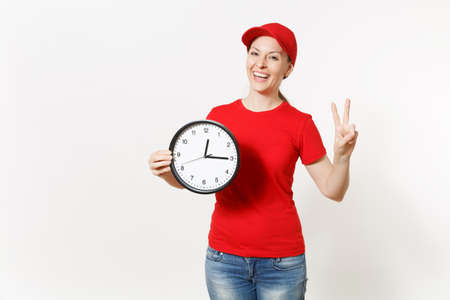 Delivery woman in red uniform isolated on white background. Professional female in cap, t-shirt, jeans working as courier or dealer, holding round clock, showing in time. Copy space for advertisementの写真素材