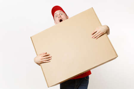 Delivery man in red uniform isolated on white background. Male in cap, t-shirt, jeans working as courier or dealer holding big large heavy cardboard box. Receiving package. Copy space advertisementの写真素材