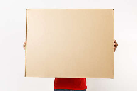 Delivery man in red uniform isolated on white background. Male in cap, t-shirt, jeans working as courier or dealer holding big large heavy cardboard box. Receiving package. Copy space advertisementの写真素材