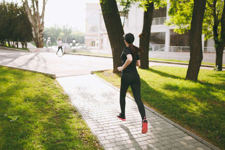Back view Young athletic brunette girl in black uniform and cap training, doing sport exercises and running, looking straight on path in city park outdoors. Fitness, healthy lifestyle conceptの写真素材