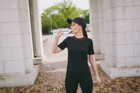 Young athletic beautiful brunette girl in black uniform and cap holding bottle, drinking water...
