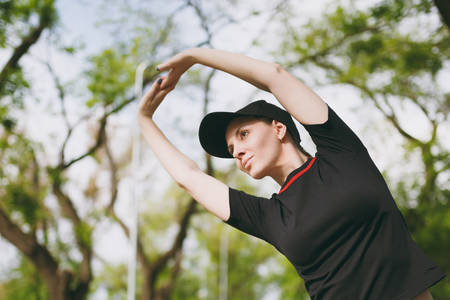 Young athletic beautiful brunette girl in black uniform, cap doing sport stretching exercises, warm-up before running or training, standing in city park outdoors. Fitness, healthy lifestyle conceptの写真素材