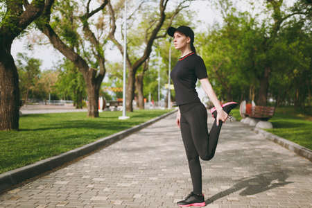 Young athletic beautiful brunette girl in black uniform, cap doing sport stretching exercises, warm-up before running or training, standing in city park outdoors. Fitness, healthy lifestyle conceptの写真素材