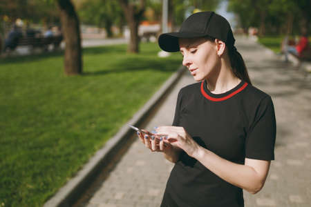 Young athletic beautiful brunette girl in black uniform and cap using mobile phone during training, looking on smartphone, standing on path in city park outdoors. Fitness, healthy lifestyle conceptの写真素材