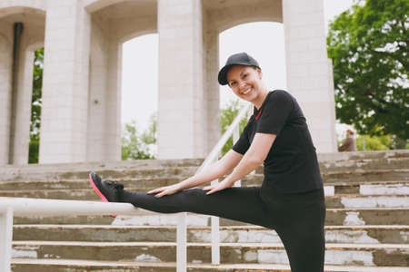 Young smiling athletic beautiful brunette girl in black uniform and cap doing sport stretching exercises, warm-up before running in city park outdoors. Fitness, healthy lifestyle conceptの写真素材