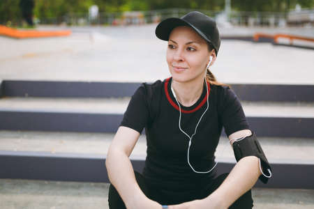 Young athletic smiling girl in black uniform and cap with headphones listening music, resting and...