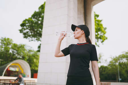 Young athletic beautiful brunette girl in black uniform and cap holding bottle, drinking water during training before running standing in city park outdoors. Fitness, healthy lifestyle conceptの写真素材