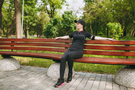 Young relaxed athletic pretty brunette girl in black uniform and cap with headphones resting after...