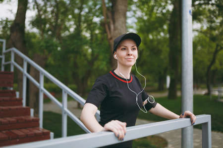 Young athletic smiling girl in black uniform and cap with headphones listening music, resting and...